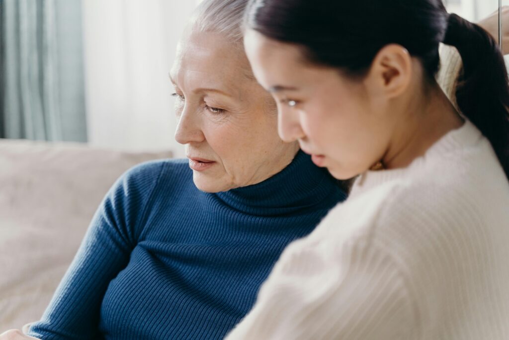 An emotional scene in a warm, modern living room showing tension between an adult daughter of color and her older mother. The daughter, appearing sad and frustrated, looks down with furrowed brows and crossed arms, while the mother stands behind her with an angry, judgmental expression. The image captures the strain and distance between them.