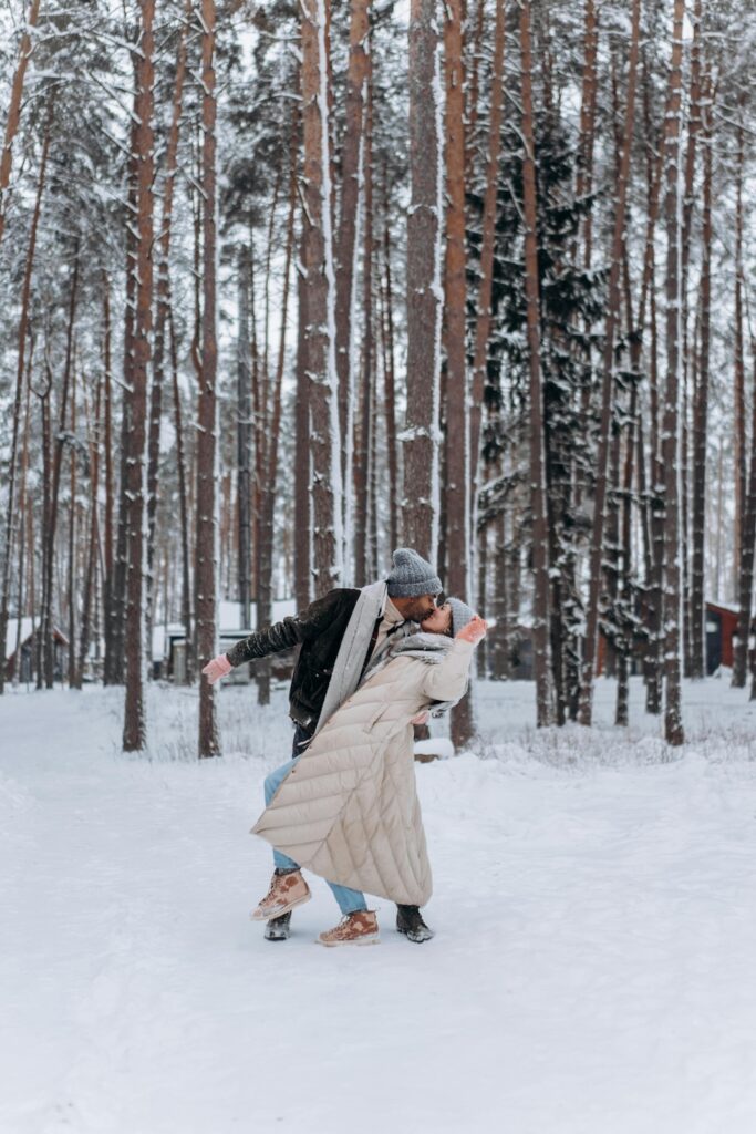 A couple shares a kiss in a snowy forest surrounded by tall pine trees. They’re bundled in winter coats, scarves, and hats, smiling as the man playfully dips the woman backward in the snow. The scene feels romantic, cozy, and joyful, capturing the warmth of connection on a cold winter day.
